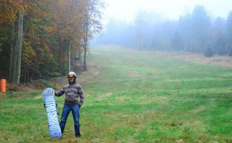 Snowboarder steht mit Ausrüstung auf Wiese, im Winter Piste, diese ist Grün, ohne Schnee, kein Spass, schaut ernst, Nitrosnowboard in der Hand, Helm und Skibrille auf